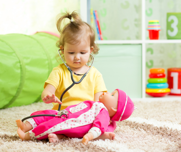 The child is playing with stethoscope, holding a doll on her knees 